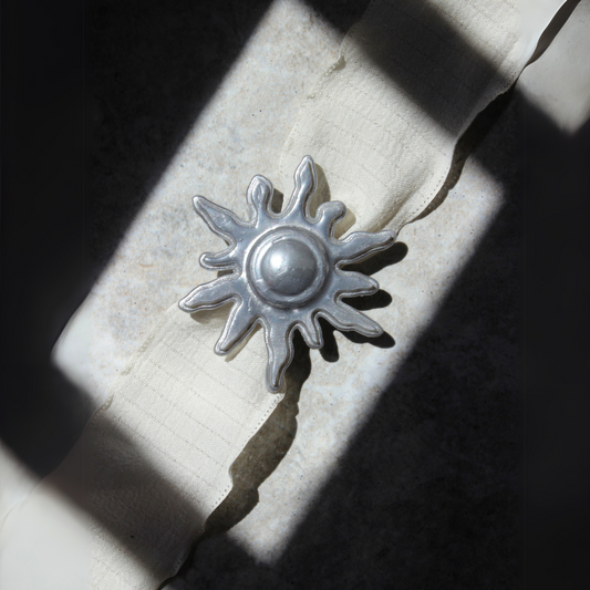 Collar artesanal con colgante de sol impreso en 3D en acabado metálico, sobre cinta de tela en color crudo.