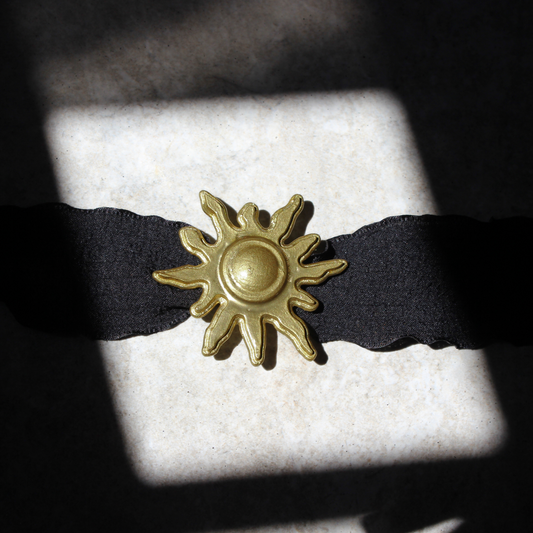 Collar artesanal con colgante de sol dorado impreso en 3D en acabado metálico, sobre cinta de tela de algodón en color negro.